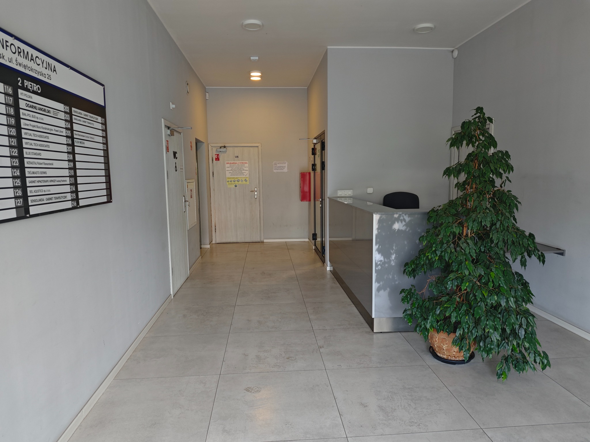 Recepcja biurowa z rośliną doniczkową i tablicą informacyjną z nazwami firm, zlokalizowana w jasnym korytarzu biurowca. Ujęcie z perspektywy wejścia.