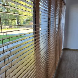 PUERTA DRZWI - Zamknięte drewniane żaluzje poziome w oknie, widok na zewnątrz z zielenią i chodnikiem.