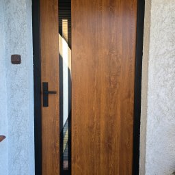 PUERTA DRZWI - Częściowo uchylone, brązowe drzwi wejściowe z czarną ramą i klamką, widok na zewnątrz przez szczelinę, obok ściana z jasnym tynkiem strukturalnym.