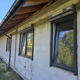 PUERTA DRZWI - Elewacja domu w trakcie budowy z zamontowanymi oknami PCV w ciemnych ramach i częściowo opuszczonymi roletami zewnętrznymi; widoczne ślady po montażu i logo producenta rolet.