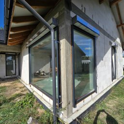 PUERTA DRZWI - Surowy stan budynku z zamontowanymi oknami z czarnymi ramami, z widocznymi roletami zewnętrznymi w kolorze szarym, instalacja orynnowania przy oknie, ściany z betonu komórkowego.