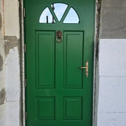 PUERTA DRZWI - Zielone drzwi zewnętrzne z przeszkleniem w górnej części, ozdobną kołatką w kształcie głowy lwa i złotą klamką, zamontowane w surowym otworze budynku w trakcie ocieplania.