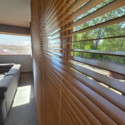 PUERTA DRZWI - Drewniane żaluzje poziome w pomieszczeniu z widokiem na zewnątrz przez okno, częściowo zasłonięte, z widocznym szarym narożnikiem kanapy i padającym światłem słonecznym na podłodze.