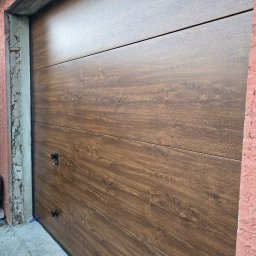 PUERTA DRZWI - Segmentowa brama garażowa o teksturze drewna, z czarnymi uchwytami i kluczem w zamku, zamontowana w surowym otworze budynku.