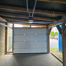 PUERTA DRZWI - Otwarta segmentowa brama garażowa w kolorze szarym, widoczne prowadnice, napęd elektryczny pod sufitem, kostka brukowa na podłodze garażu, dwa samochody i tuje w tle.