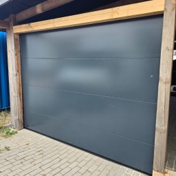 PUERTA DRZWI - Szara, segmentowa brama garażowa z drewnianą konstrukcją nośną, widok z zewnątrz na kostkę brukową.
