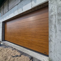 PUERTA DRZWI - Nowa brama garażowa segmentowa w kolorze drewna, zamontowana w surowym otoczeniu budowy w miejscowości Pisz, widoczna z bliska na tle betonowych ścian.
