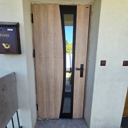 PUERTA DRZWI - Drewnopodobne drzwi wejściowe z czarną, asymetryczną wstawką i przeszkleniem, widok na zewnątrz, skrzynka pocztowa po lewej stronie.