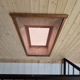 PUERTA DRZWI - Drewniane okno dachowe z roletą wewnętrzną, widok od dołu, wkomponowane w drewniany sufit z boazerii, fragment metalowej barierki w tle.