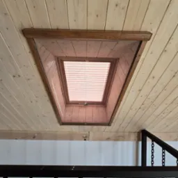 Drewniane okno dachowe z roletą wewnętrzną, widok od dołu, wkomponowane w drewniany sufit z boazerii, fragment metalowej barierki w tle.