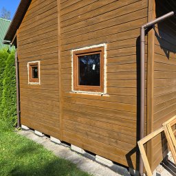 PUERTA DRZWI - Montaż okna w drewnianym domu, widoczne pianowanie wokół ramy, obok leżą stare ramy okienne.