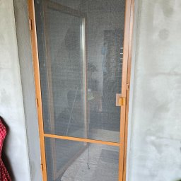 PUERTA DRZWI - Moskitiera w drewnianej ramie, zamontowana w surowym otworze drzwiowym z widocznym tynkiem, widok na wnętrze pomieszczenia przez siatkę.