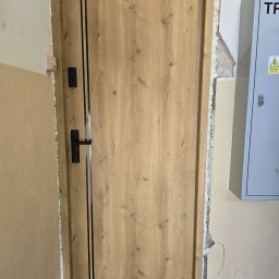 PUERTA DRZWI - Montaż brązowych drzwi wewnętrznych z czarnym zamkiem i pionowymi paskami, widoczne wypełnienie pianką montażową w szczelinie ościeżnicy, obok skrzynka elektryczna.