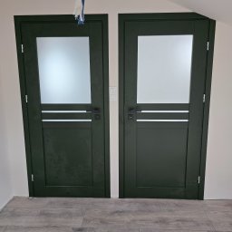 PUERTA DRZWI - Dwie ciemnozielone drzwi wewnętrzne z matowym szkłem, wstawka z trzema poziomymi paskami, czarne klamki, widoczne ślady montażu przy podłodze.
