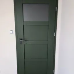 Zielone drzwi wewnętrzne z szybą w górnej części, zamontowane w nowym budynku z widocznym gniazdkiem elektrycznym bez obudowy i surową podłogą.