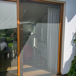 PUERTA DRZWI - Zewnętrzna plisowana moskitiera w brązowej ramie zamontowana na drzwiach tarasowych, odbicie fotografa w szybie, biała elewacja z zielonymi pnączami.