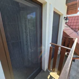 Zewnętrzna plisa moskitierowa w brązowej ramie, zamontowana na balkonie, widoczne rury instalacyjne na elewacji i fragment czerwonej dachówki w tle.