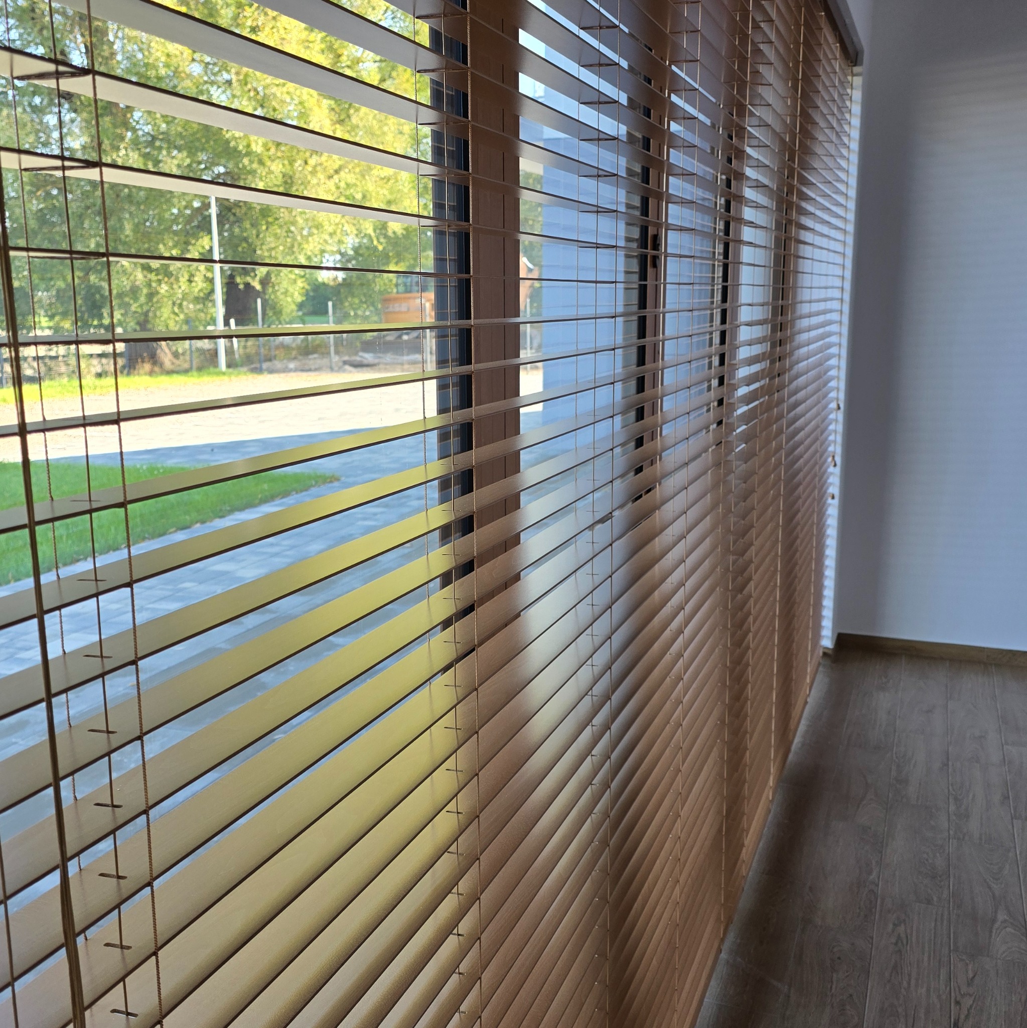 Zamknięte drewniane żaluzje poziome w oknie, widok na zewnątrz z zielenią i chodnikiem.