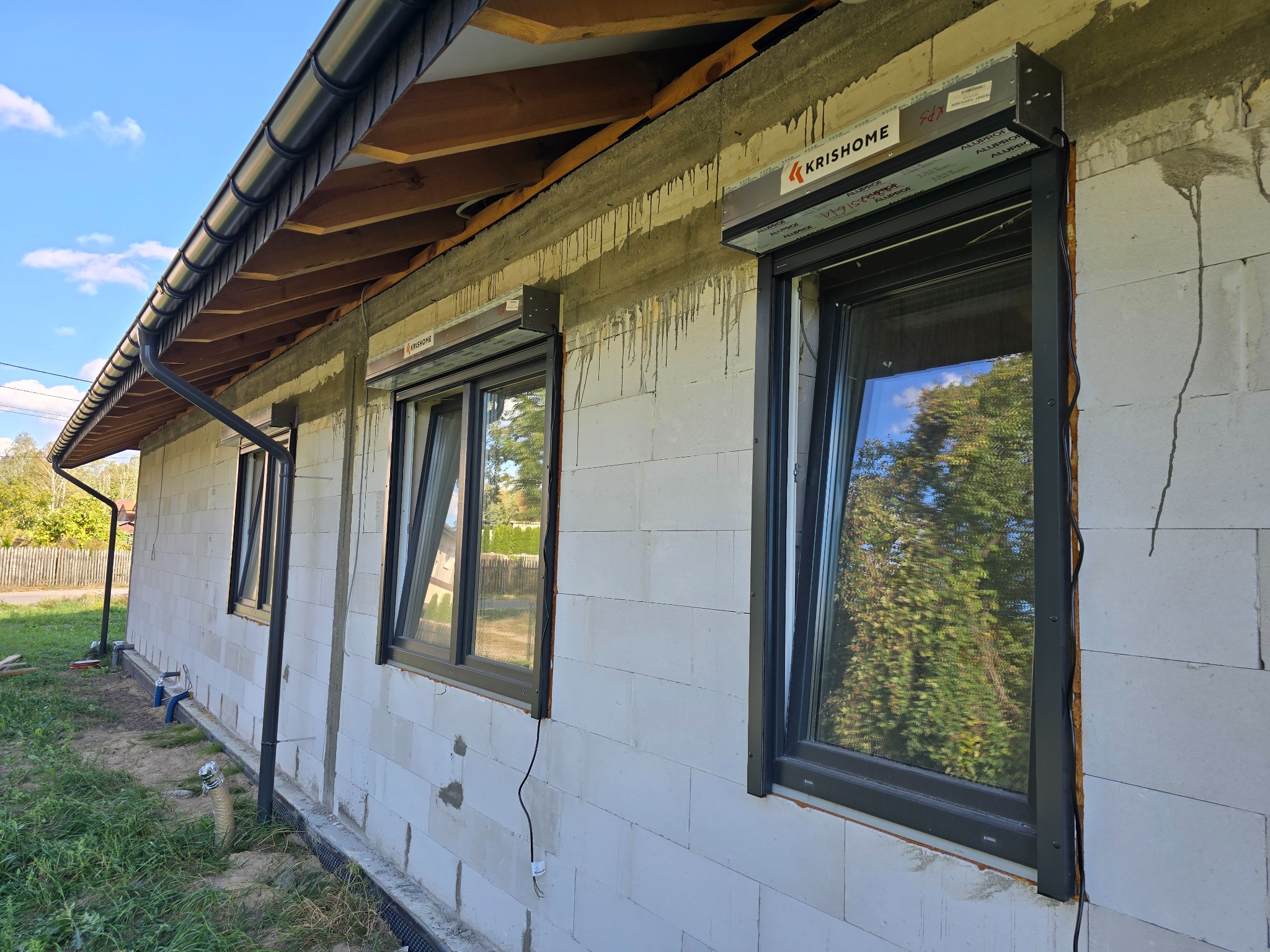 Elewacja domu w trakcie budowy z zamontowanymi oknami PCV w ciemnych ramach i częściowo opuszczonymi roletami zewnętrznymi; widoczne ślady po montażu i logo producenta rolet.