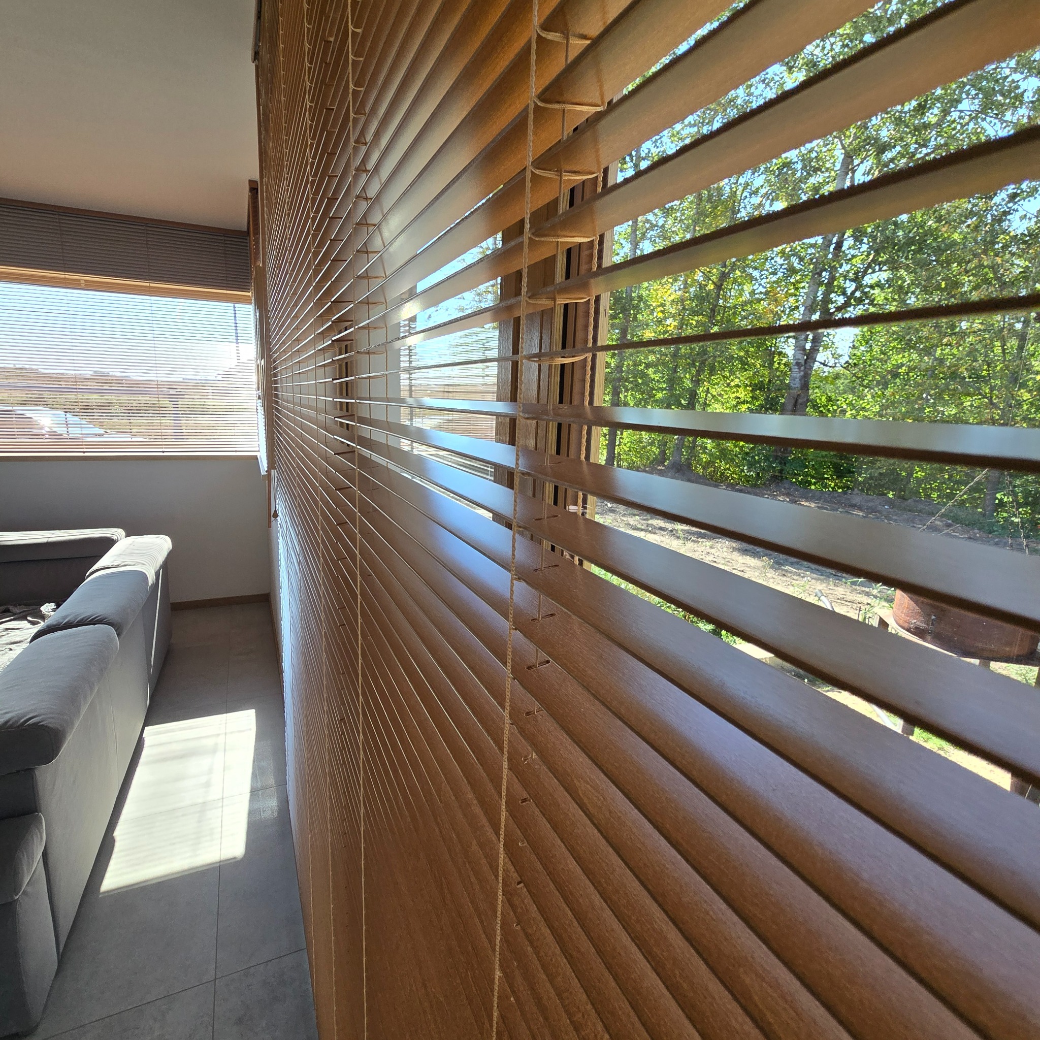 Drewniane żaluzje poziome w pomieszczeniu z widokiem na zewnątrz przez okno, częściowo zasłonięte, z widocznym szarym narożnikiem kanapy i padającym światłem słonecznym na podłodze.