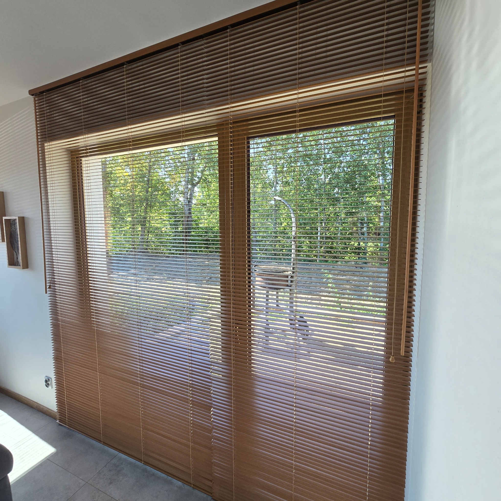 Brązowe żaluzje poziome zamontowane w oknie, widok na zieleń drzew za oknem, jasne wnętrze.