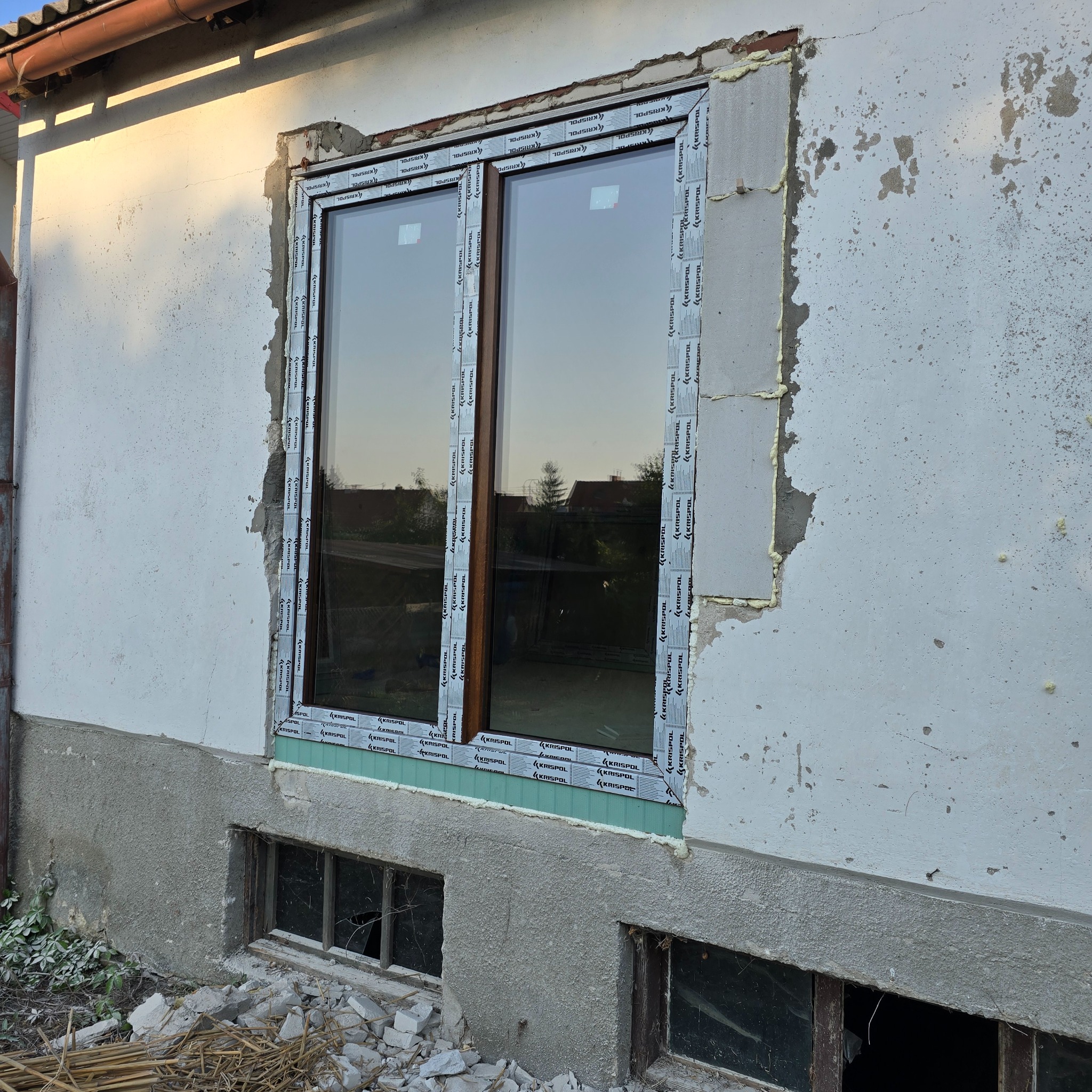 Montaż nowego okna dwuszybowego z brązowymi ramami w surowym stanie, widoczne pianka montażowa, niedokończone wykończenie elewacji i stare okna piwniczne poniżej.
