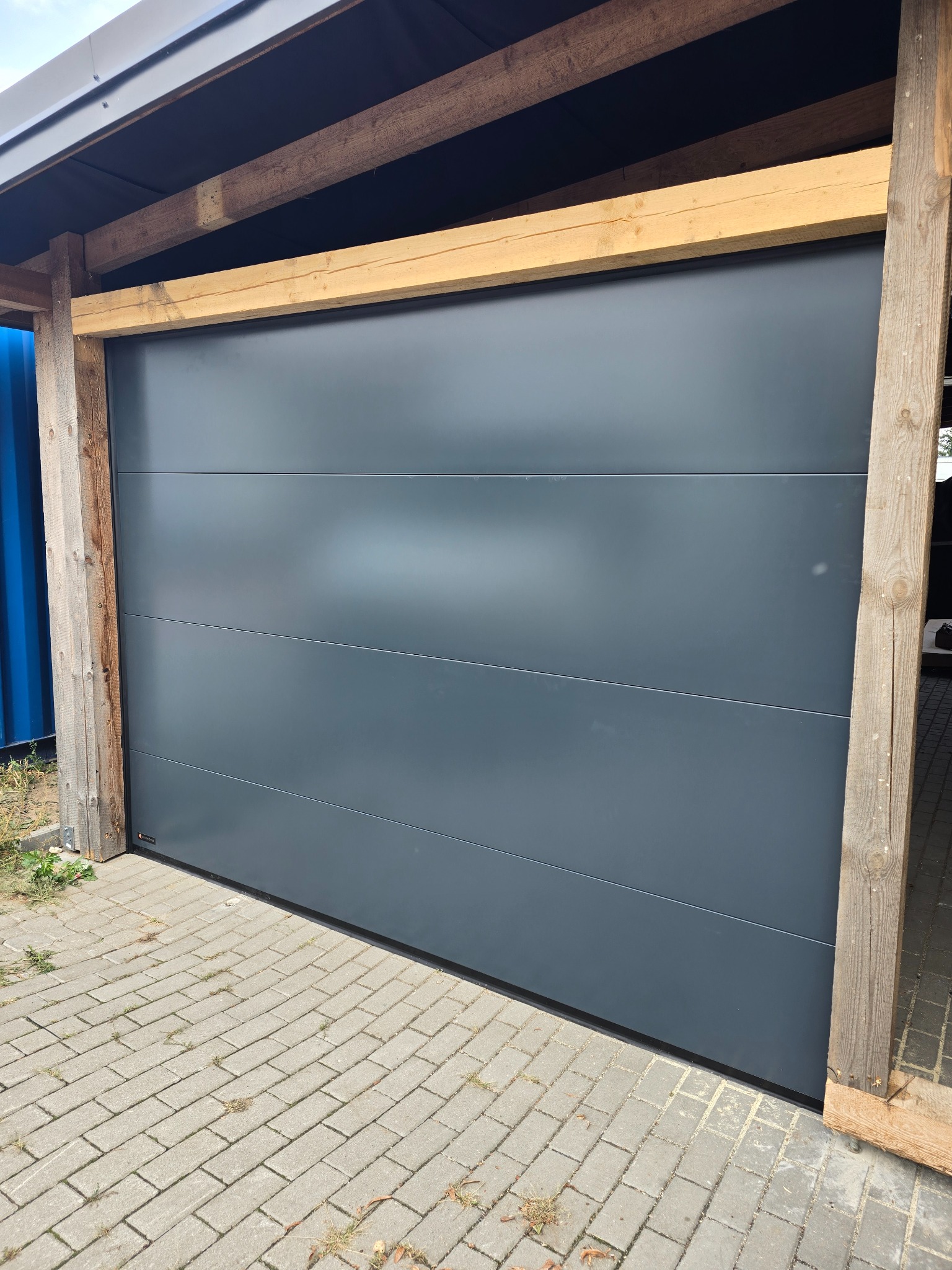 Szara, segmentowa brama garażowa z drewnianą konstrukcją nośną, widok z zewnątrz na kostkę brukową.