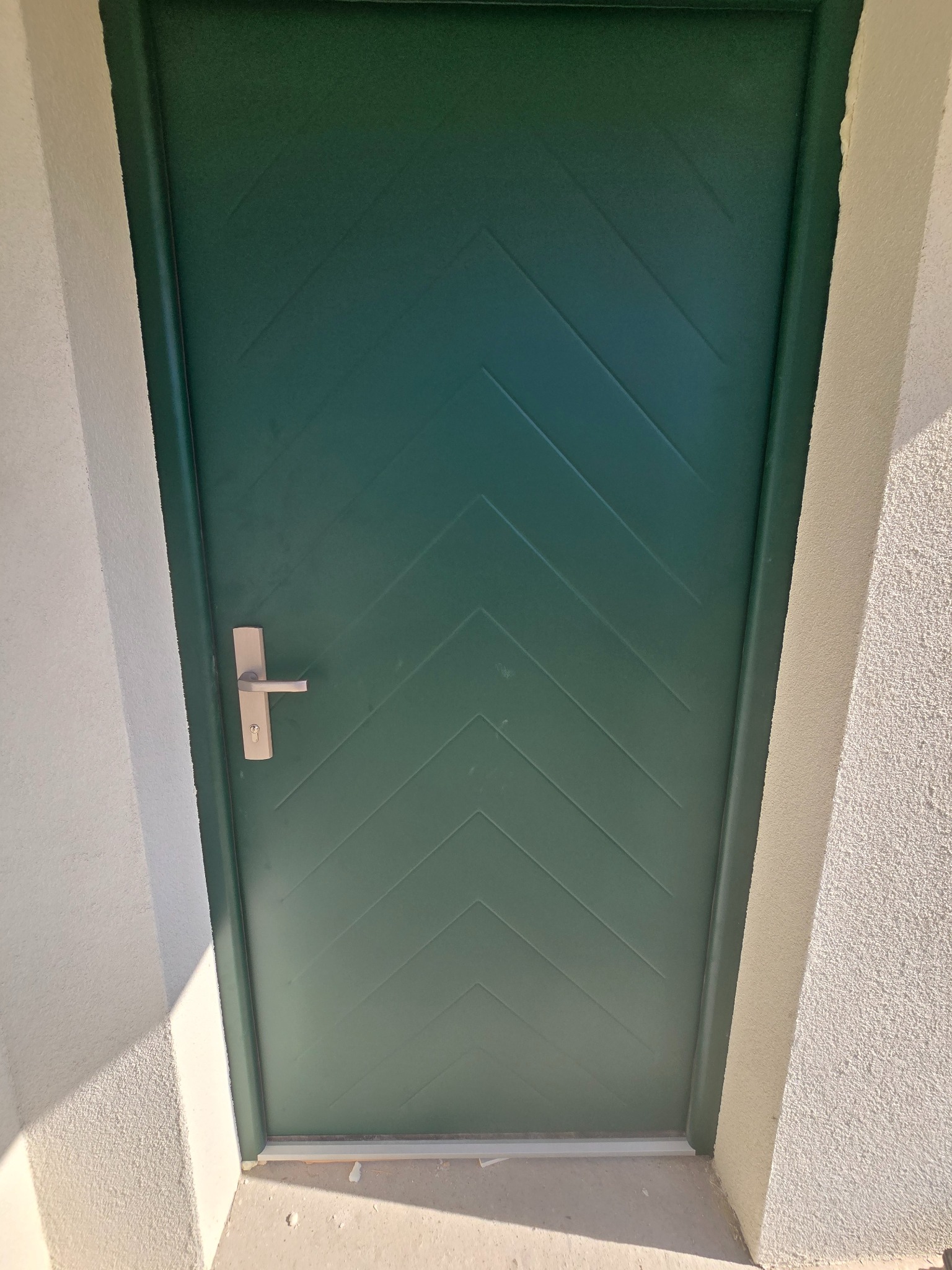 Zielone drzwi zewnętrzne z poziomym wzorem w jodełkę i srebrną klamką, zamontowane w białej ścianie z fakturą.