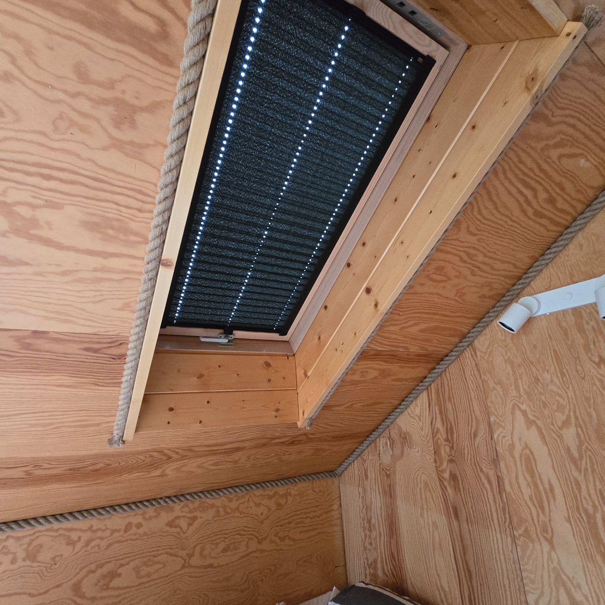 Czarna plisowana roleta okienna z białymi diodami LED, zamontowana w drewnianej ramie okna dachowego, otoczona dekoracyjnym sznurem jutowym.