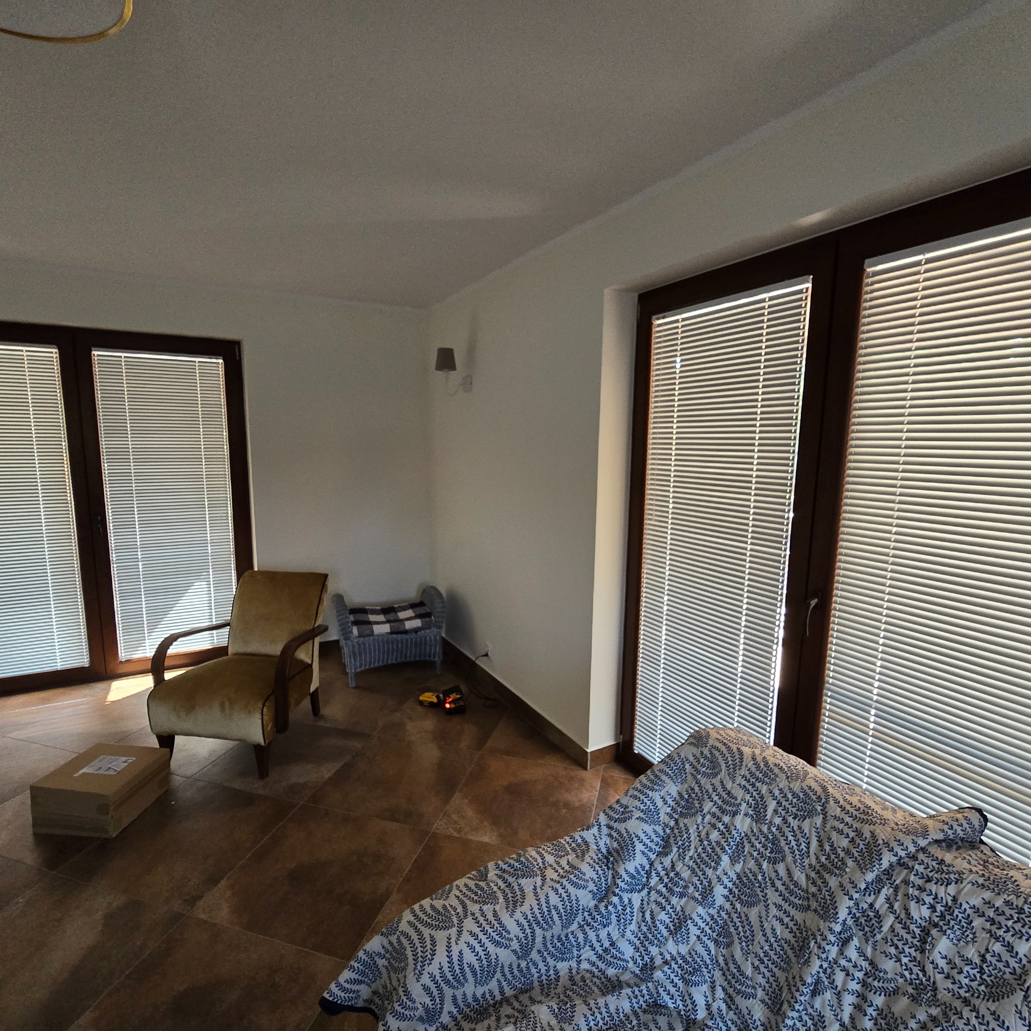 Wnętrze pokoju z brązowymi oknami z żaluzjami, fotel w stylu retro, karton na podłodze i niebieska narzuta na kanapie.