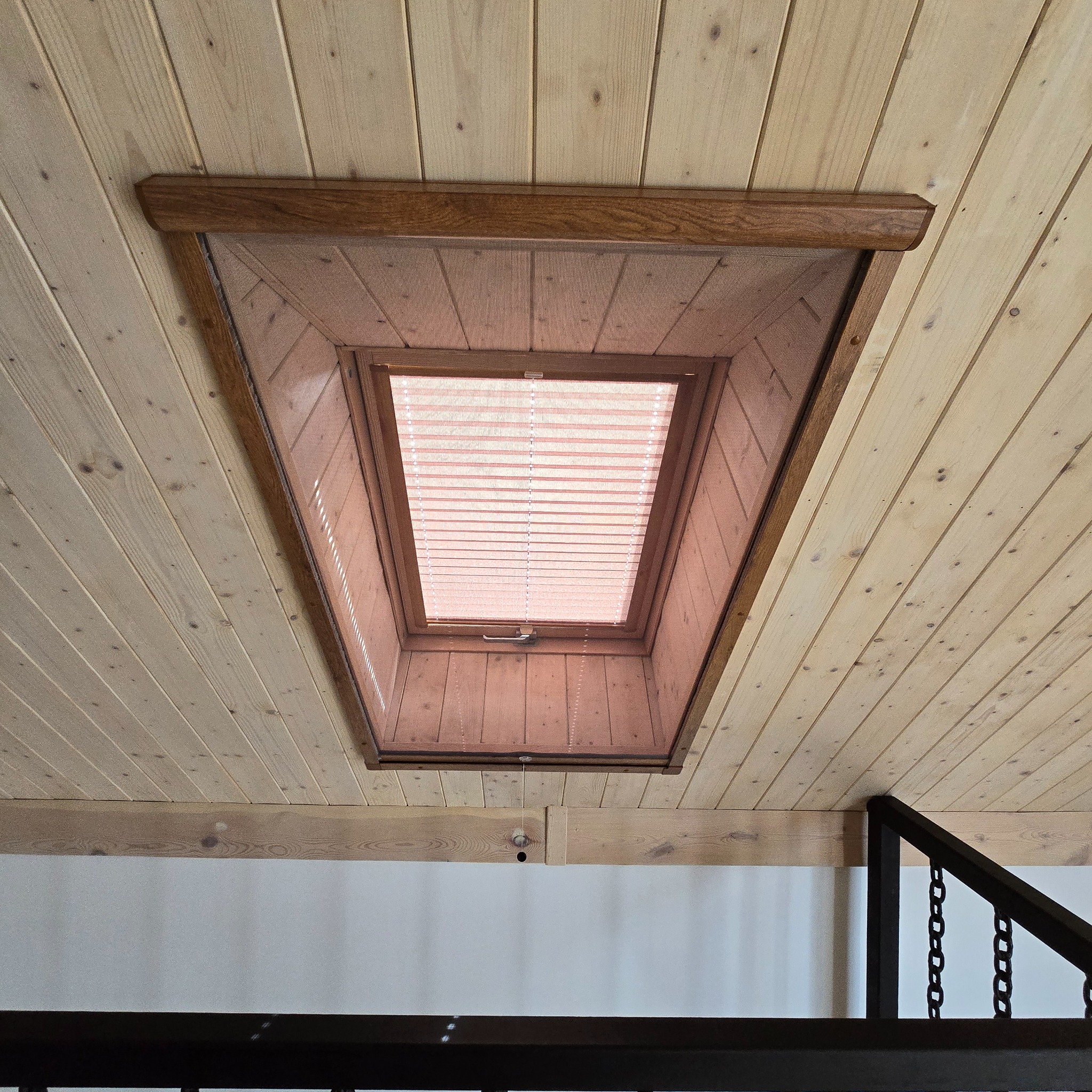 Drewniane okno dachowe z roletą wewnętrzną, widok od dołu, wkomponowane w drewniany sufit z boazerii, fragment metalowej barierki w tle.