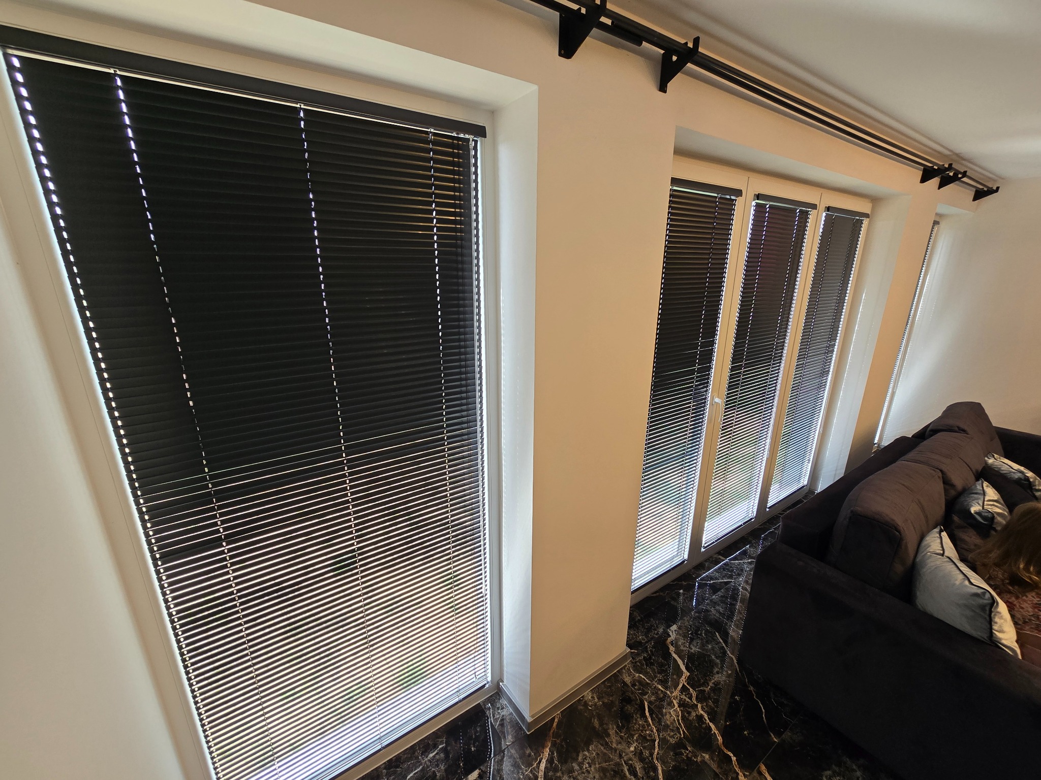 Czarne żaluzje poziome zamontowane w oknach, widok z dołu, marmurowa podłoga, fragment sofy w tle.