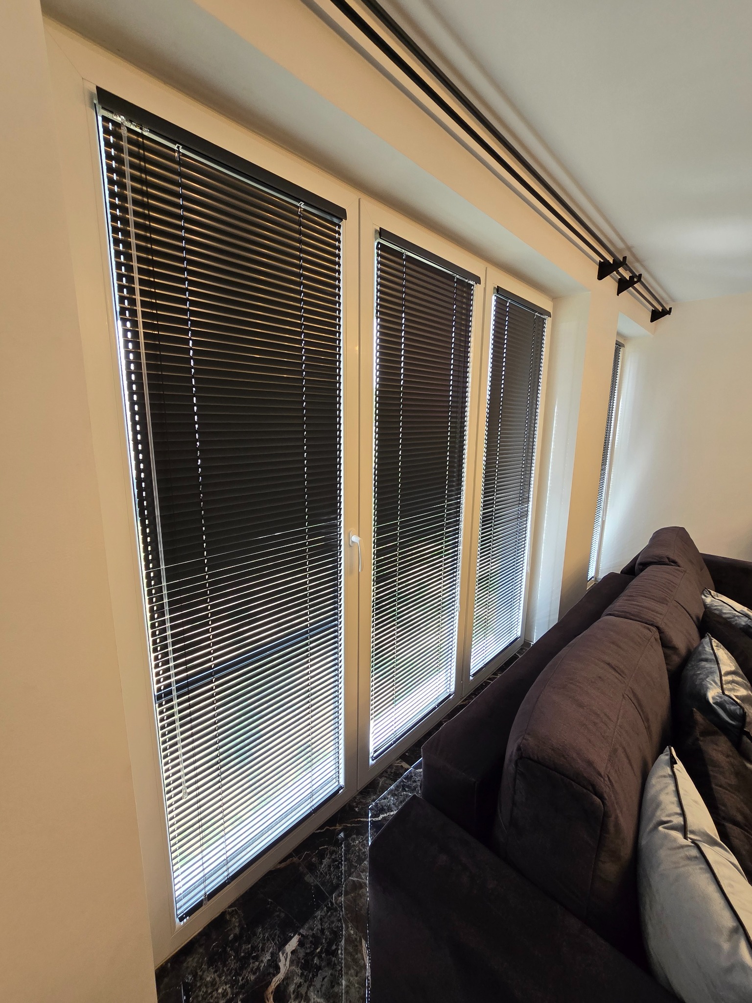 Trzy okna z białymi ramami i zamontowanymi ciemnymi żaluzjami poziomymi, widok z wnętrza salonu z fragmentem sofy.