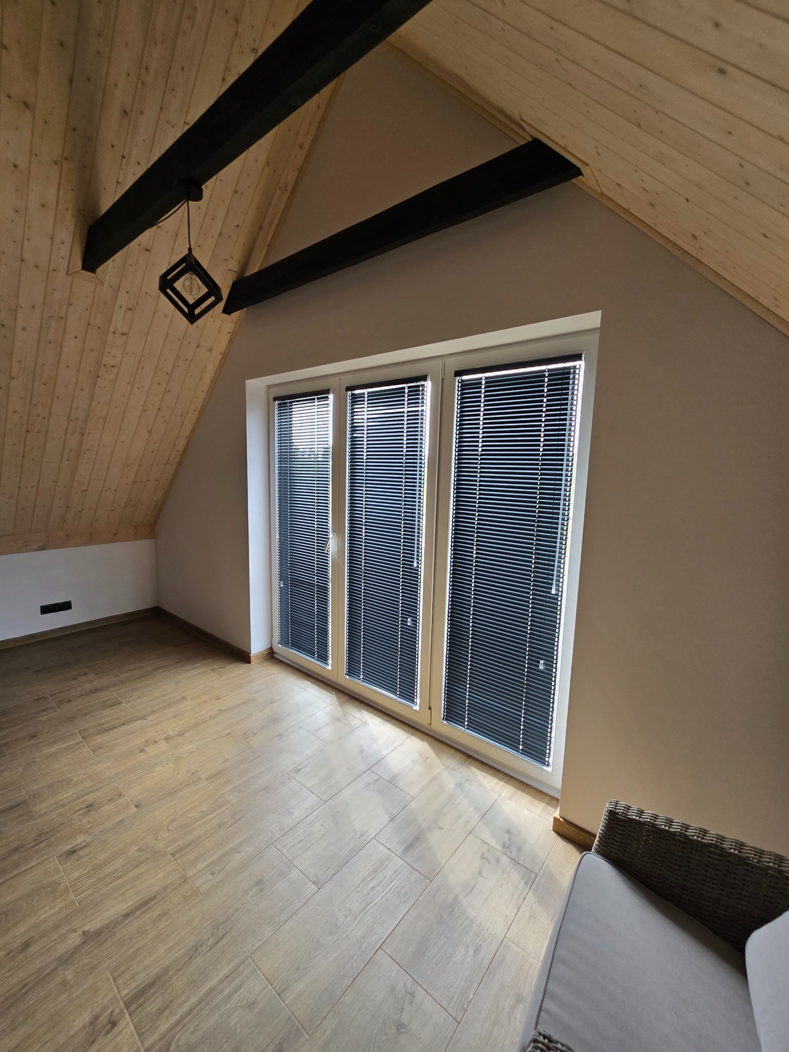 Poddasze z drewnianym stropem i trzema oknami z żaluzjami poziomymi, widok z dołu na skos dachu, nowoczesna lampa na czarnej belce stropowej.