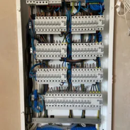 Rozdzielnica elektryczna z zamontowanymi bezpiecznikami i przewodami, widok z bliska na kompletną instalację w białej obudowie.