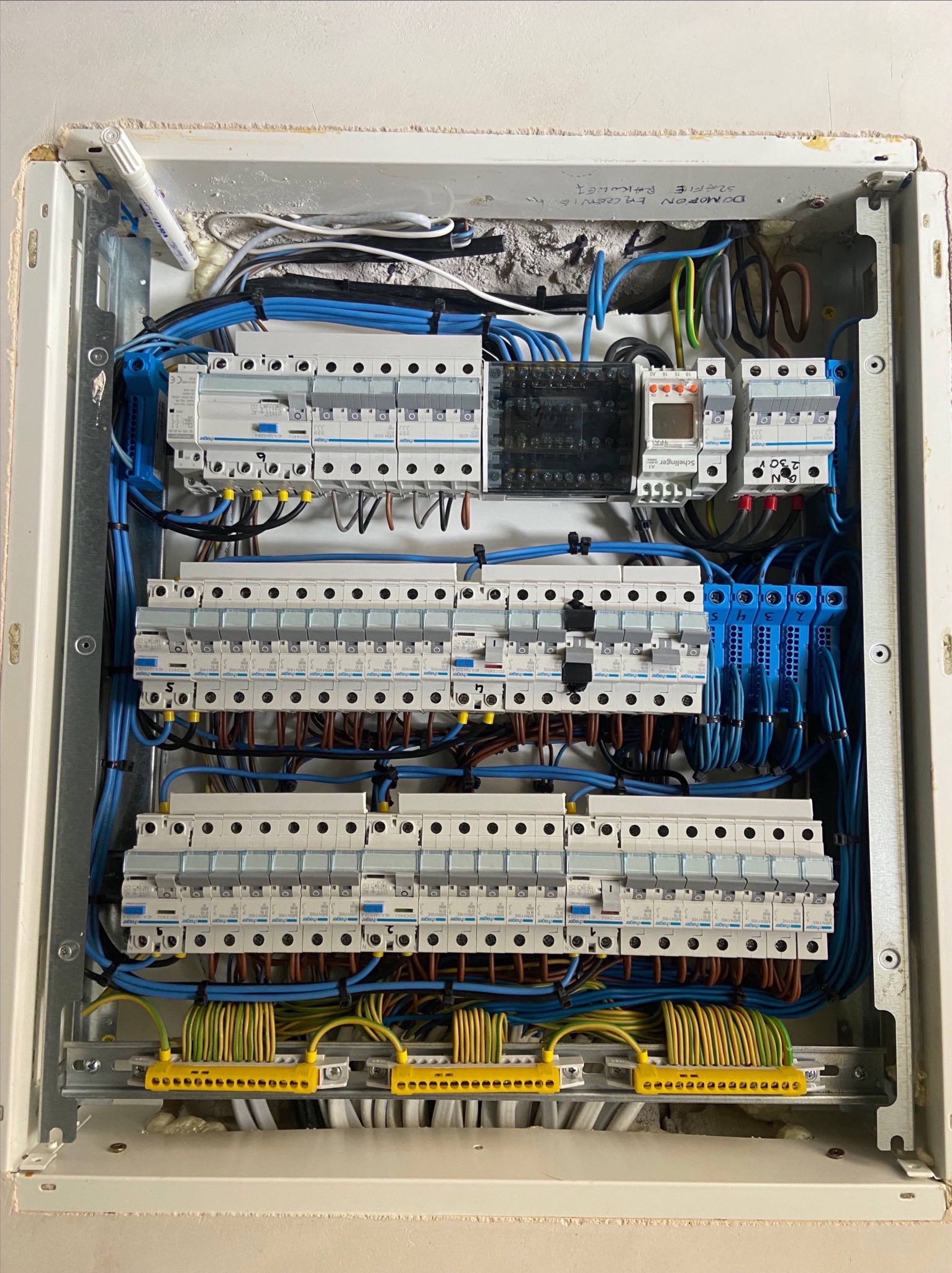 Otwarta skrzynka elektryczna z zainstalowanymi bezpiecznikami, okablowaniem w kolorach niebieskim, brązowym, żółto-zielonym i czarnym, widoczne oznaczenia na obudowie i elementach, ślady pianki...