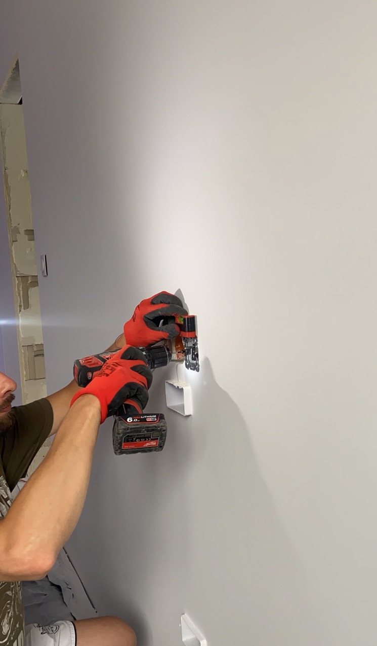 Elektryk w czerwonych rękawicach montuje gniazdko elektryczne w szarej ścianie za pomocą wiertarki akumulatorowej Milwaukee, widoczny fragment twarzy i fragment niedokończonego remontu w tle.