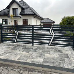 Loftico - Nowoczesna, pozioma brama wjazdowa z geometrycznym wzorem w kształcie strzał, prowadząca do domu z panelami słonecznymi na dachu garażu, widoczna z poziomu kostki brukowej.