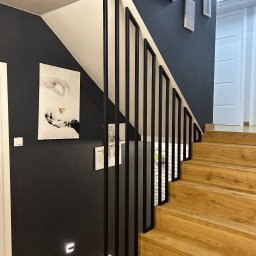 Loftico - Nowoczesne schody z drewnianymi stopniami i czarną metalową balustradą, oświetlenie punktowe w ścianie, obrazy na ścianie w tle.