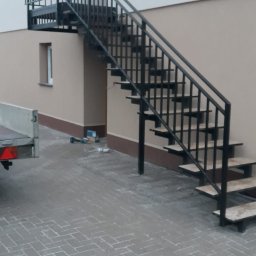 Loftico - Zewnętrzne schody z czarną metalową konstrukcją i drewnianymi stopniami, prowadzące na balkon, widoczne narzędzia i przyczepka samochodowa w tle.