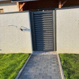 Loftico - Nowoczesna, grafitowa brama wjazdowa z poziomymi lamelami, umieszczona w jasnym, otynkowanym murze z widocznym okablowaniem, prowadząca do domu z panelami słonecznymi na dachu, otoczona trawnikiem...