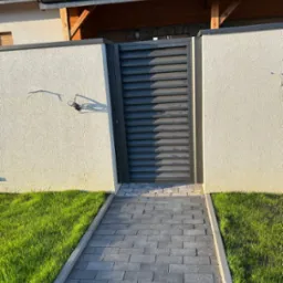 Nowoczesna, grafitowa brama wjazdowa z poziomymi lamelami, umieszczona w jasnym, otynkowanym murze z widocznym okablowaniem, prowadząca do domu z panelami słonecznymi na dachu, otoczona trawnikiem...