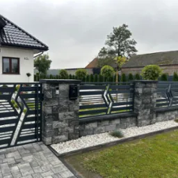 Nowoczesne, grafitowe ogrodzenie panelowe z bramą wjazdową, z elementami dekoracyjnymi w kształcie rombów, osadzone na kamiennych słupkach, przed domem z czarną dachówką, w tle zabudowania...