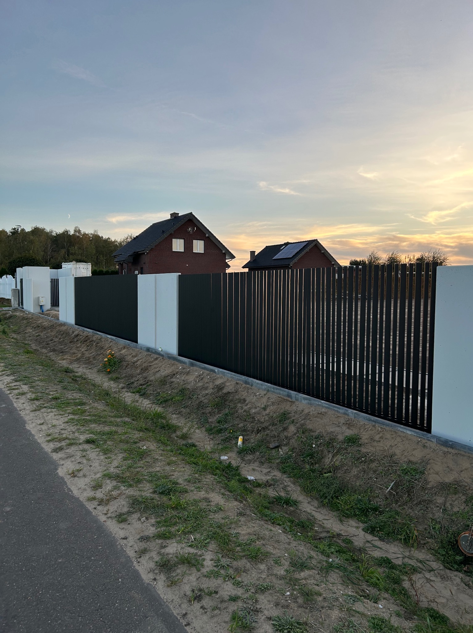 Nowoczesne ogrodzenie panelowe z pionowych sztachet w kolorze antracytowym, osadzone na betonowych słupkach, na tle domów jednorodzinnych o zachodzie słońca.