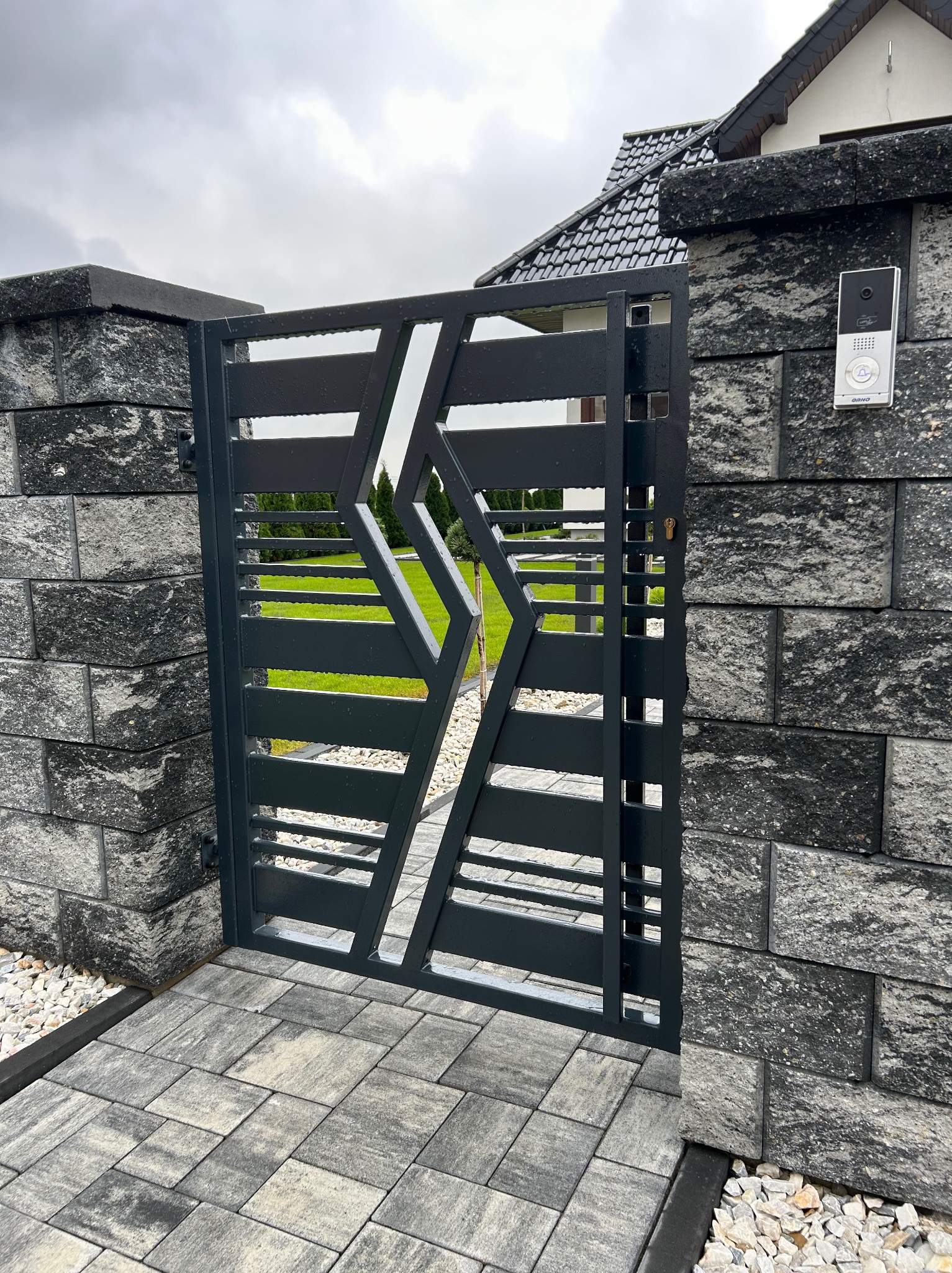 Nowoczesna, grafitowa brama wjazdowa z poziomymi szczeblami i geometrycznym wzorem rombu, osadzona między dwoma słupkami z szarej, łupanej kostki, z widocznym domofonem po prawej stronie i kostką...