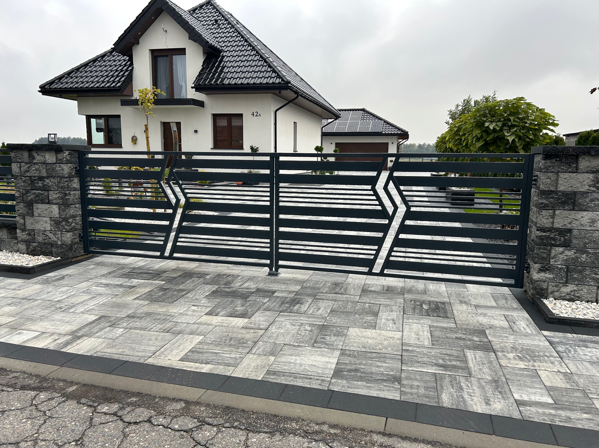 Nowoczesna, pozioma brama wjazdowa z geometrycznym wzorem w kształcie strzał, prowadząca do domu z panelami słonecznymi na dachu garażu, widoczna z poziomu kostki brukowej.