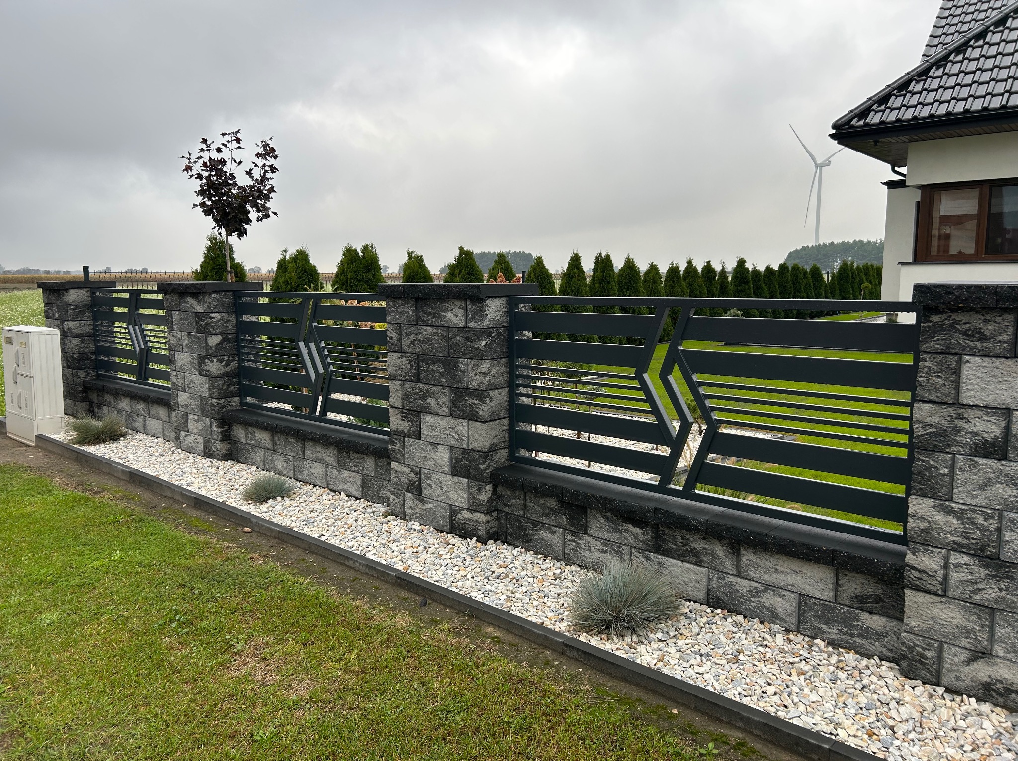 Nowoczesne ogrodzenie panelowe z elementami kamiennymi w kolorze grafitowym, ozdobione białym żwirem i roślinami, na tle domu z turbiną wiatrową.