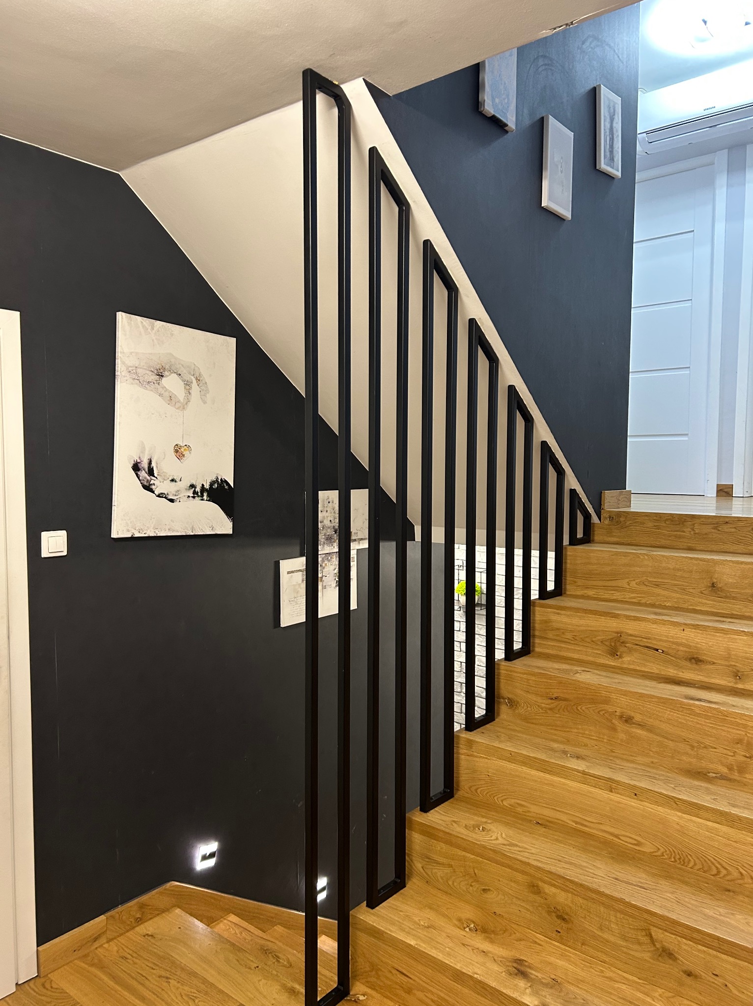 Nowoczesne schody z drewnianymi stopniami i czarną metalową balustradą, oświetlenie punktowe w ścianie, obrazy na ścianie w tle.