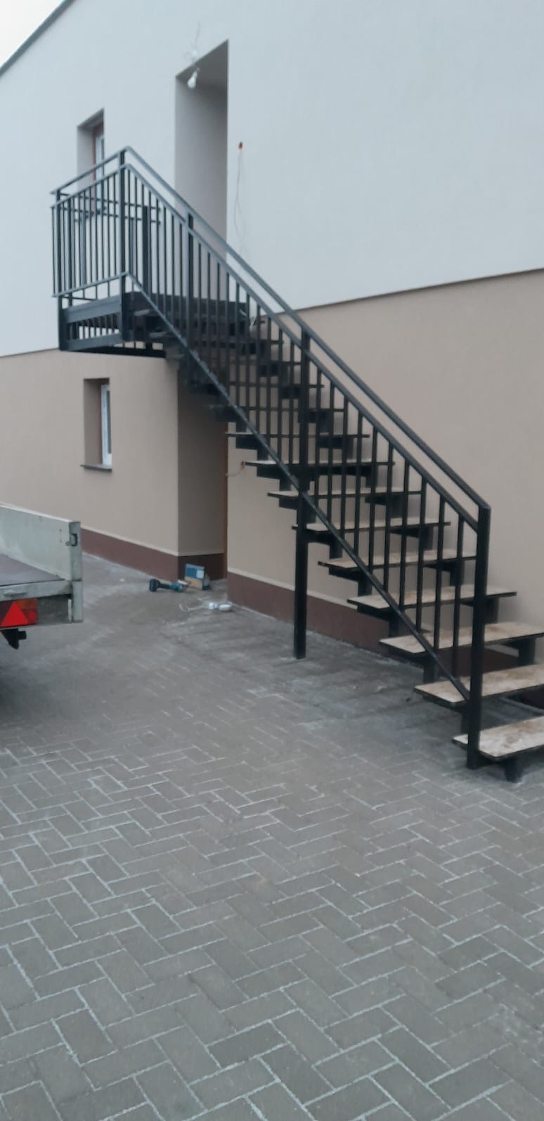 Zewnętrzne schody z czarną metalową konstrukcją i drewnianymi stopniami, prowadzące na balkon, widoczne narzędzia i przyczepka samochodowa w tle.