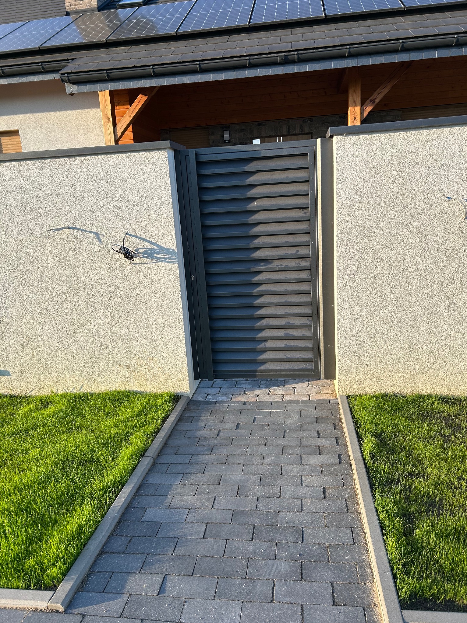 Nowoczesna, grafitowa brama wjazdowa z poziomymi lamelami, umieszczona w jasnym, otynkowanym murze z widocznym okablowaniem, prowadząca do domu z panelami słonecznymi na dachu, otoczona trawnikiem...